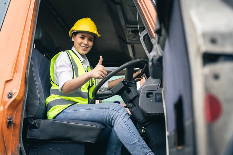 A smiling woman wearing a yellow hard hat, white shirt, and high-visibility vest sits in the driver's seat of an LTL Freight Truck